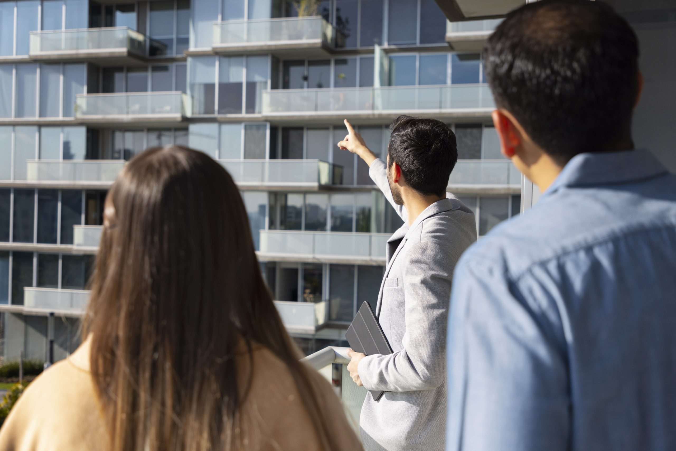 Group of people on a balcony looking at a modern glass apartment building; one man points toward the building while others observe.